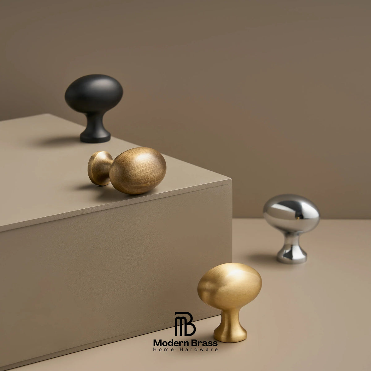Four modern round cabinet knobs in matte black, brushed gold, satin silver, and polished gold, displayed on a beige pedestal and surface against a warm taupe background. The Modern Brass Home Hardware logo is visible at the bottom.