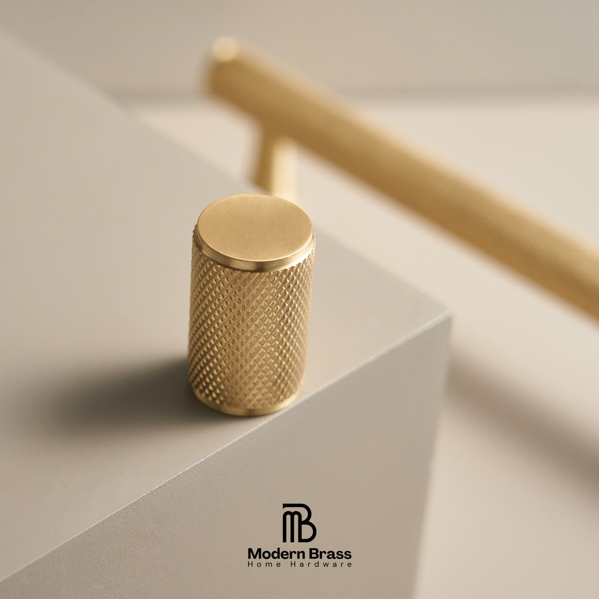 A close-up of a cylindrical brass cabinet knob with a knurled textured surface, resting on a light gray geometric block. A larger, out-of-focus brass knob is visible in the background.