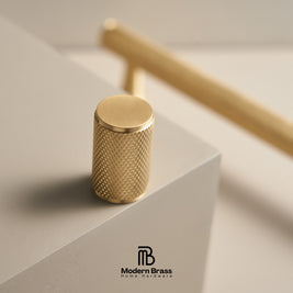 A close-up of a cylindrical brass cabinet knob with a knurled textured surface, resting on a light gray geometric block. A larger, out-of-focus brass knob is visible in the background.