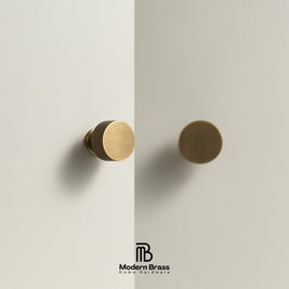 Two round cabinet knobs in a brushed brass finish, one with a knurled side and one smooth, mounted on a light beige wall or door. The Modern Brass Home Hardware logo is visible at the bottom.