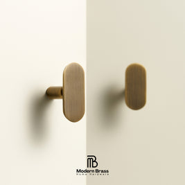 Two oval brass cabinet knobs mounted on a light beige door, with a vertical seam down the center and a Modern Brass logo at the bottom.