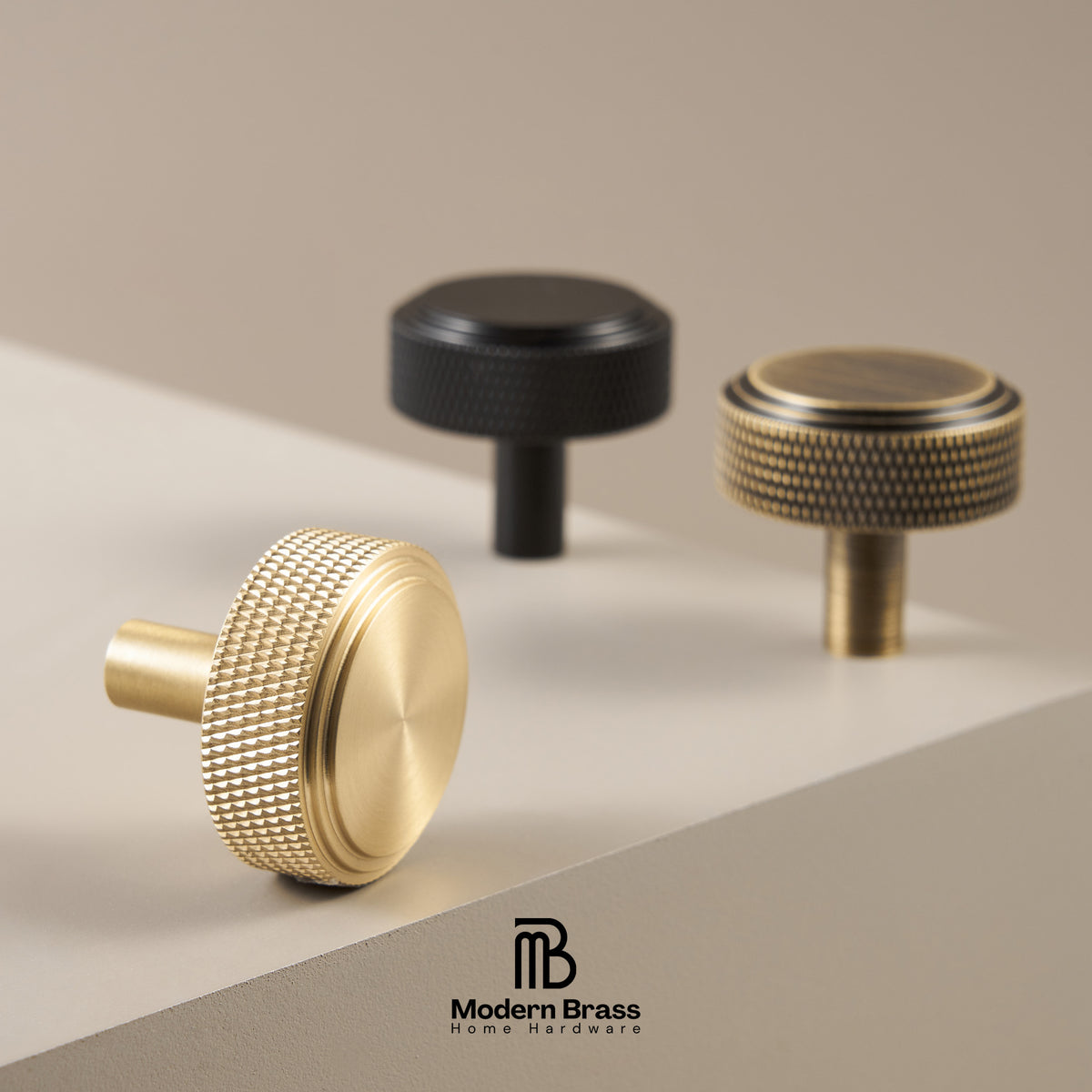 Three brass cabinet knobs in different finishes (matte gold, textured gold, and matte black) arranged on a light beige surface with a neutral background. The Modern Brass logo is visible at the bottom.