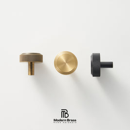 Three round cabinet knobs in black, matte brass, and brushed brass are arranged on a light beige surface against a warm beige background. The Modern Brass logo is visible at the bottom.