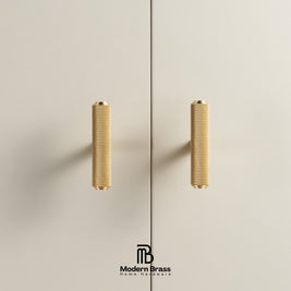 Two modern cylindrical brass cabinet handles with a textured knurled finish, mounted on a light beige wall or door. A vertical seam runs down the center, and the 'Modern Brass' brand logo is visible at the bottom.