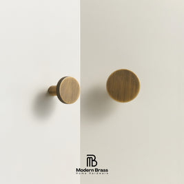 Two round brass cabinet knobs in a warm brushed finish, mounted on a clean off-white wall with a vertical seam running down the center. The Modern Brass logo appears at the bottom.