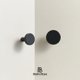 Two round cabinet knobs in matte black finishes shown mounted on a light beige wall, with the Modern Brass Home Hardware logo at the bottom.
