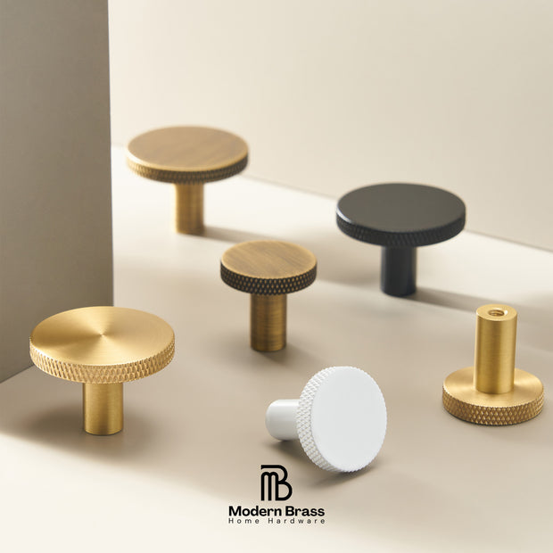 Six modern cabinet knobs in various finishes (brass, black, white) arranged on a minimalist beige surface with a geometric background. The Modern Brass logo is visible at the bottom.