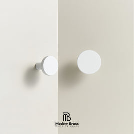 Two white round cabinet knobs mounted on a light beige wall, with one knocker featuring a textured outer ring and the other a smooth matte finish. The Modern Brass Home Hardware logo is visible at the bottom.