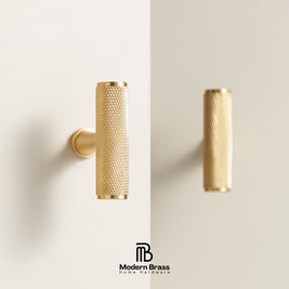 A modern cylindrical brass wall hook with a textured mesh pattern, mounted on a clean off-white wall. A reflection of the hook is visible on the right side of the wall, and the 'Modern Brass Home Hardware' logo is displayed at the bottom.