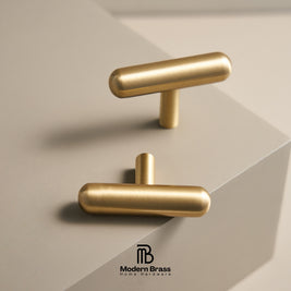 Two modern T-shaped cabinet knobs in a brushed brass finish, displayed on a minimalist light beige geometric pedestal. The Modern Brass logo is visible at the bottom.