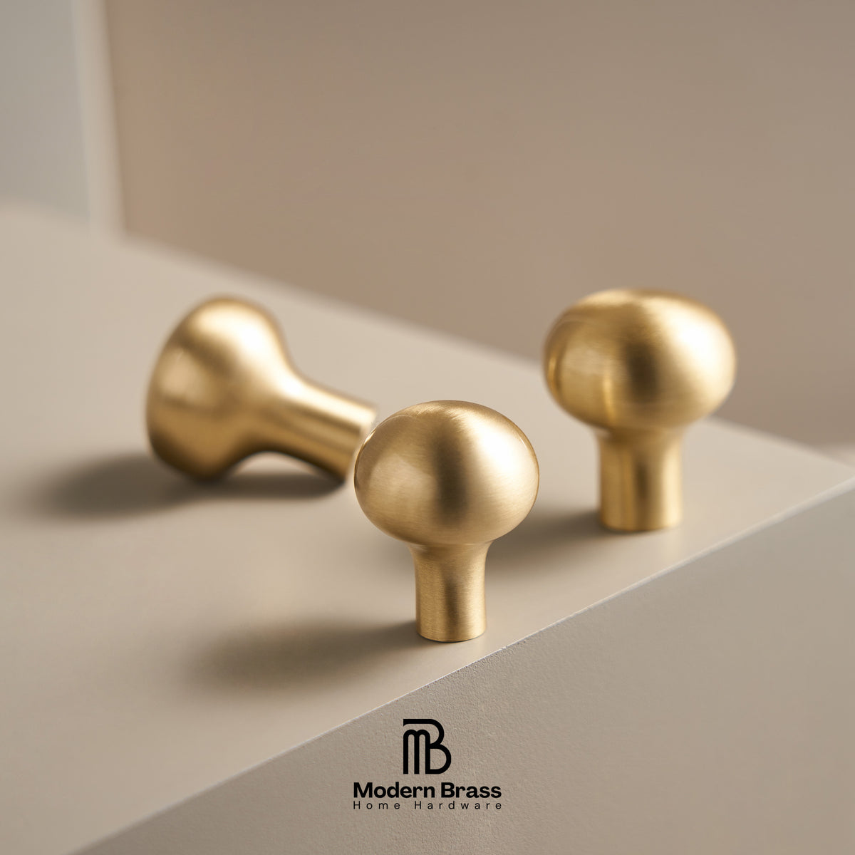 Three brass cabinet knobs in different shapes, including a round ball knob and two mushroom-shaped knobs, displayed on a light beige surface with a soft shadow. The Modern Brass logo is visible at the bottom.