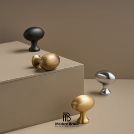 Four modern round cabinet knobs in matte black, brushed gold, satin silver, and polished gold, displayed on a beige pedestal and surface against a warm taupe background. The Modern Brass Home Hardware logo is visible at the bottom.