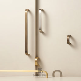 Modern brass and bronze cabinet handles mounted on a light beige wall, including long vertical bar handles, a smaller curved pull, and a top-mounted pull, with a Modern Brass logo at the bottom.