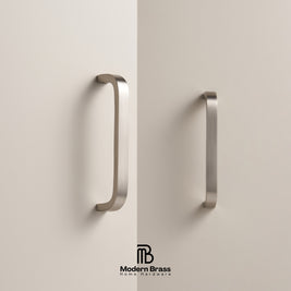 Two modern curved cabinet handles in a brushed brass finish mounted on a light beige door, with the Modern Brass Home Hardware logo at the bottom.
