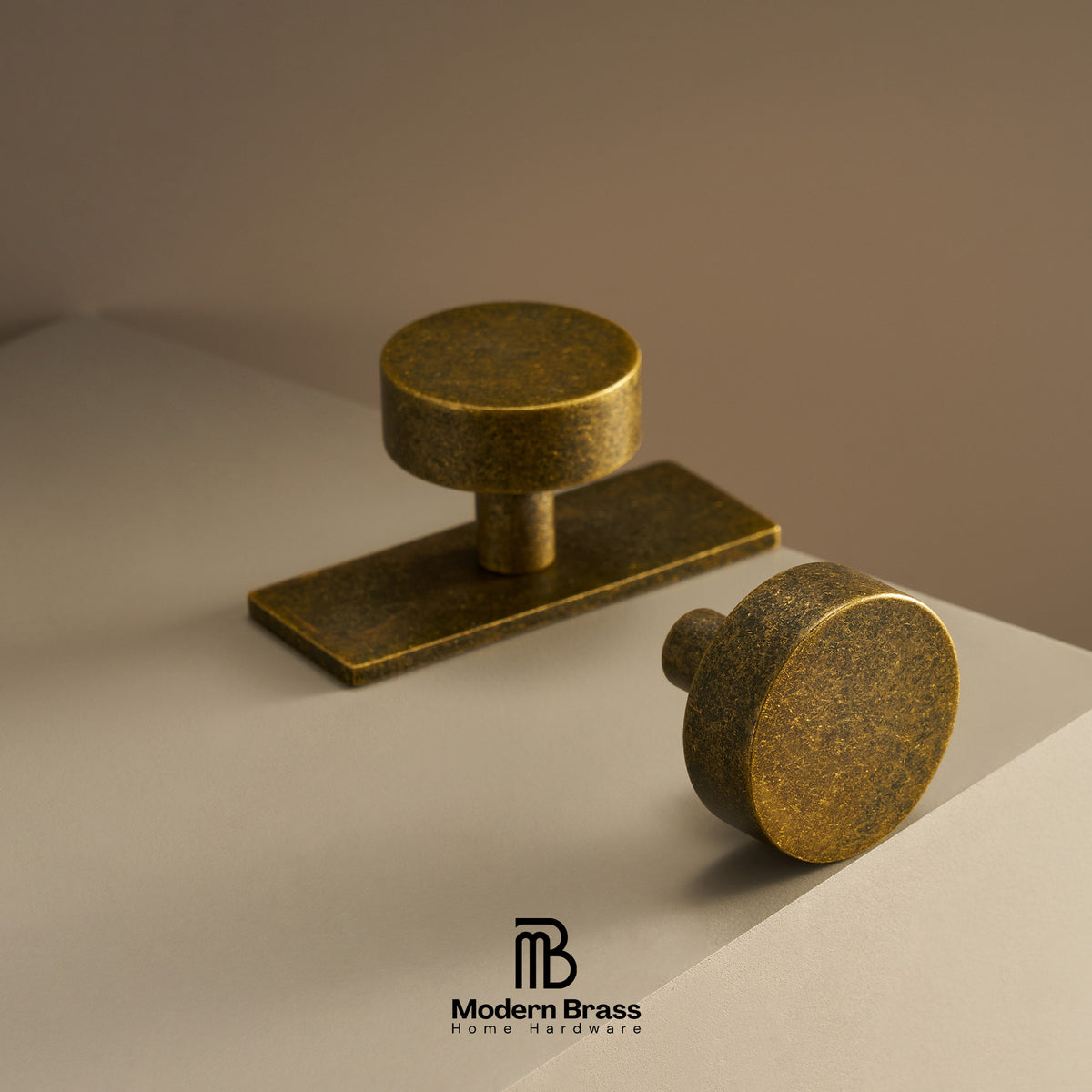 Two antique brass cabinet knobs, one rectangular pull and one round knob, displayed on a neutral beige surface with a matching background. The Modern Brass logo is visible at the bottom.