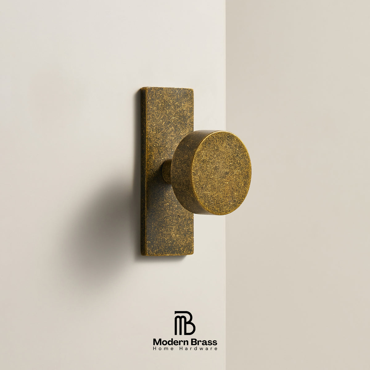 A round brass cabinet knob with a rectangular backplate, mounted on a light beige wall. The Modern Brass logo is visible at the bottom.