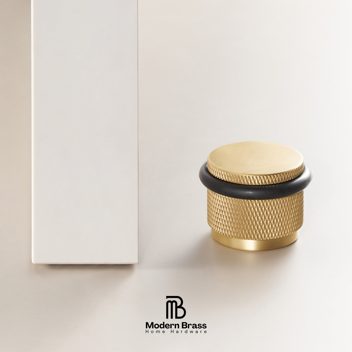A brass cylindrical home hardware knob with a textured knurled pattern, a flat circular top, and a black rubber accent ring, shown next to its white packaging box on a light beige background. The Modern Brass logo is visible at the bottom.