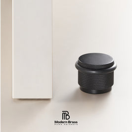 Three round cabinet knobs in black, matte black, and brushed brass are displayed on a light beige surface against a warm beige background. The Modern Brass logo is visible at the bottom.
