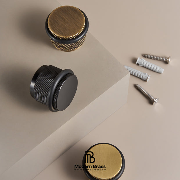 Three round cabinet knobs in brushed brass and matte black finishes, shown with mounting screws and wall anchors on a beige geometric studio background. A Modern Brass logo watermark appears near the bottom.