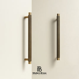 Two long cylindrical cabinet handles in a brushed brass finish with gold-toned end caps, mounted on a light beige wall. The Modern Brass logo is visible at the bottom.