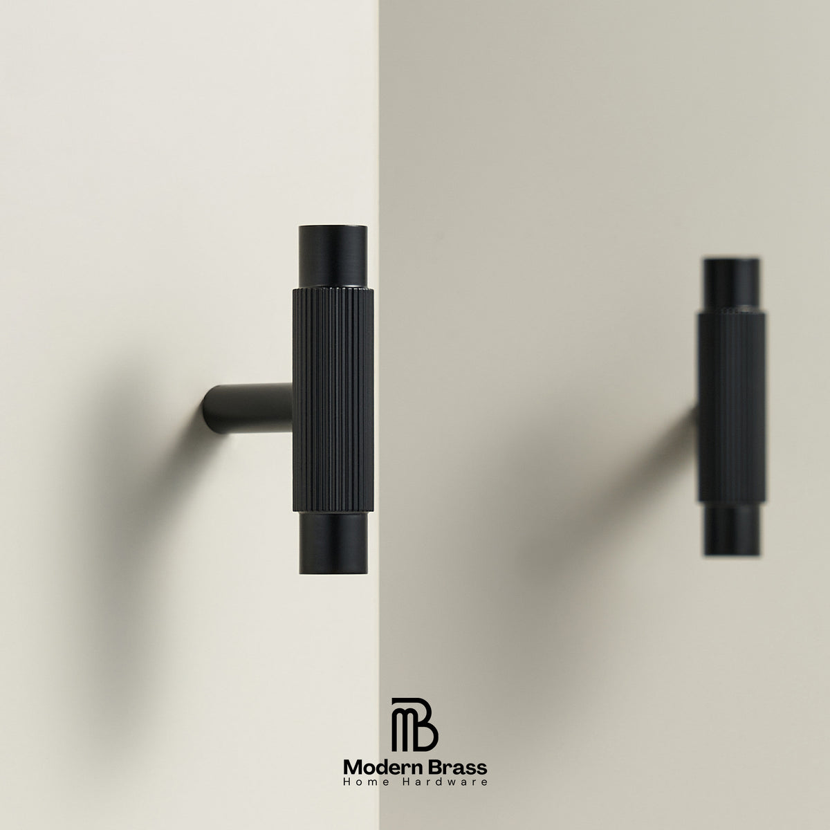 Two matte black wall-mounted cabinet knobs with a ribbed cylindrical design, shown on a split beige background with the Modern Brass Home Hardware logo at the bottom.