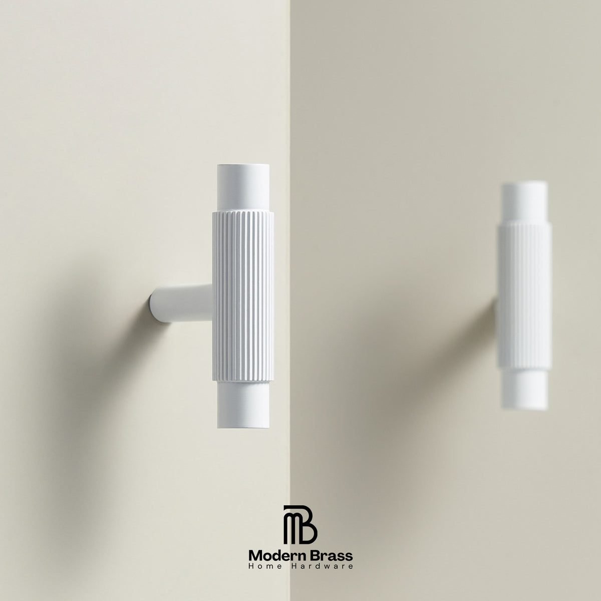 A pair of modern white wall-mounted brass hooks with ribbed cylindrical handles, shown on a warm beige wall with a reflection effect and the Modern Brass logo at the bottom.