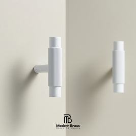 A pair of modern white wall-mounted brass hooks with ribbed cylindrical handles, shown on a warm beige wall with a reflection effect and the Modern Brass logo at the bottom.