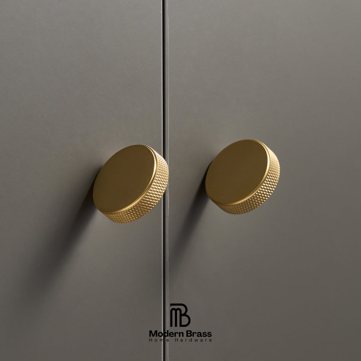 Two round brushed brass cabinet knobs with a knurled textured edge, mounted on matte gray doors. The Modern Brass logo is visible at the bottom.