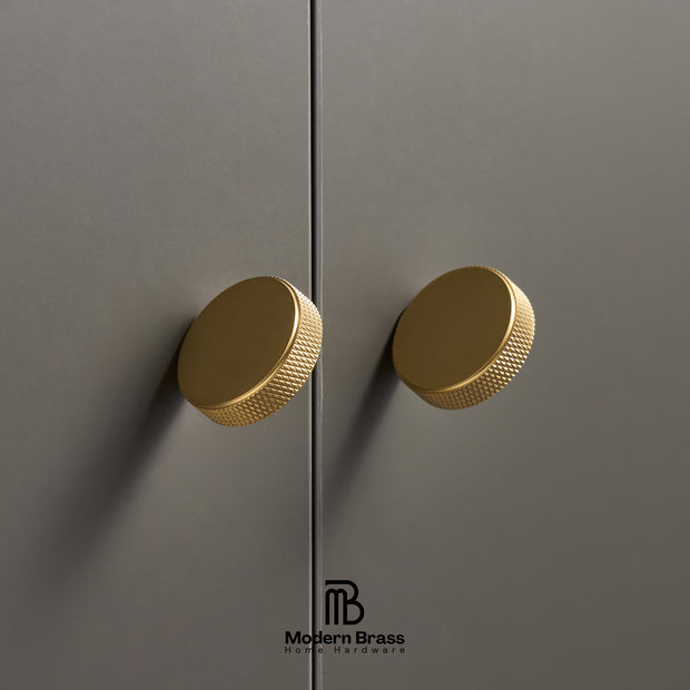Two round brushed brass cabinet knobs with a knurled textured edge, mounted on matte gray doors. The Modern Brass logo is visible at the bottom.
