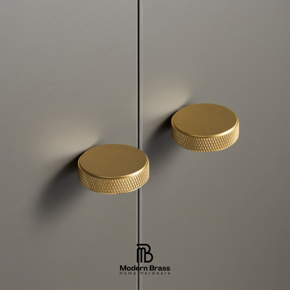 Two round brushed brass cabinet pulls with a knurled textured edge, mounted on a matte gray door with a vertical seam running down the center. The Modern Brass logo is visible at the bottom.