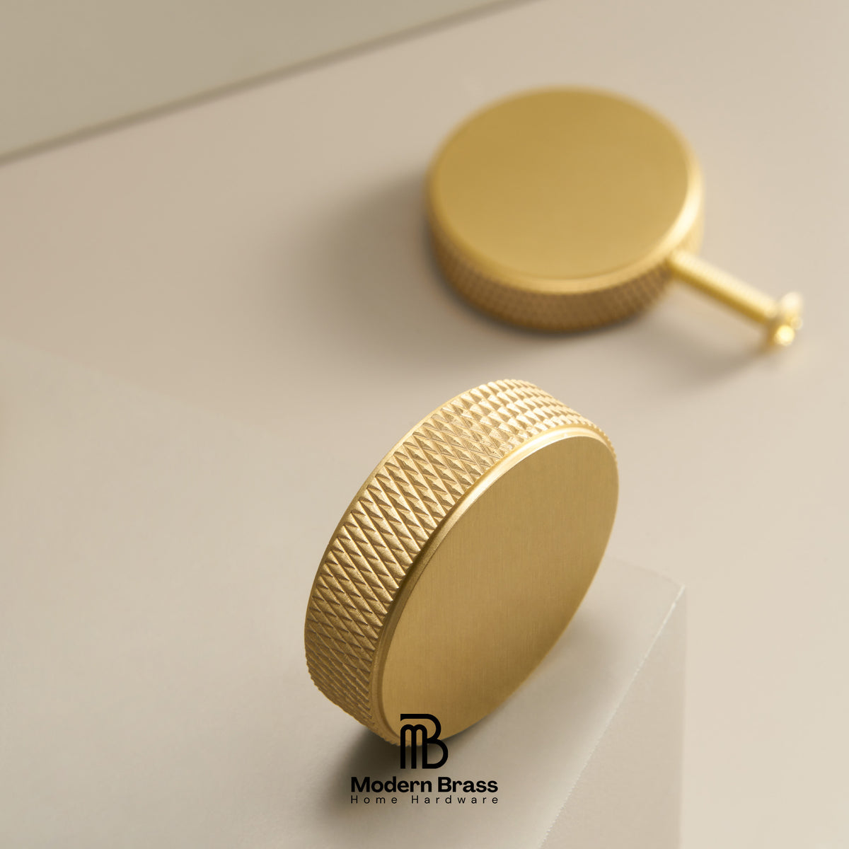 Two round gold-toned hardware knobs with a brushed finish and knurled textured edges, shown on a clean light beige surface. One knob is upright in the foreground and the other lies on its side in the background.