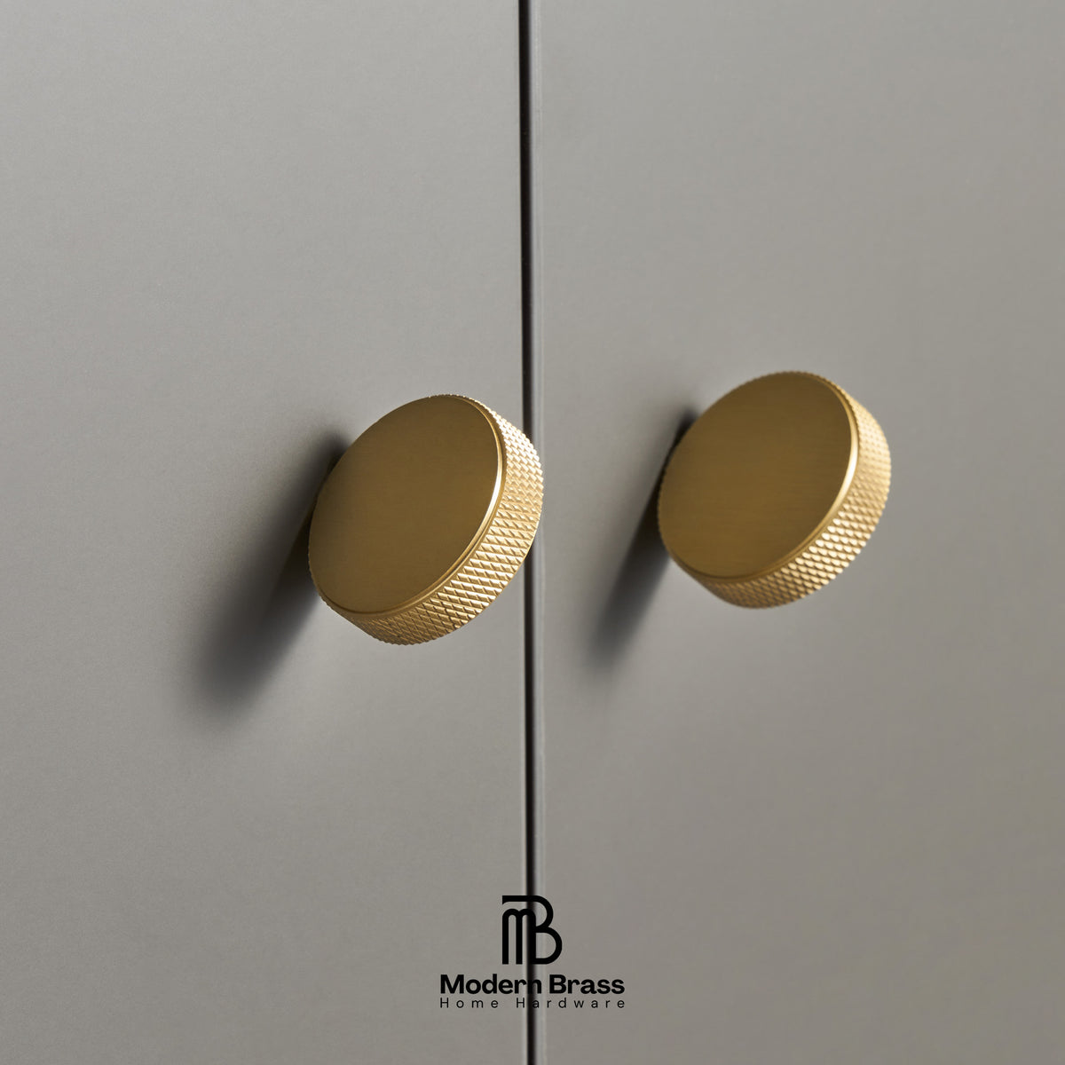 Two round brushed brass cabinet knobs mounted on a gray door, with a Modern Brass logo at the bottom.