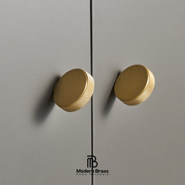 Two round brushed brass cabinet knobs mounted on a gray door, with a Modern Brass logo at the bottom.