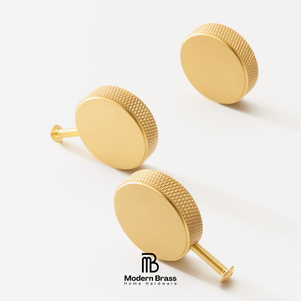 Three round gold cabinet knobs with a textured outer edge and flat front face, shown on a clean white background. One knob is turned to show the threaded back post.