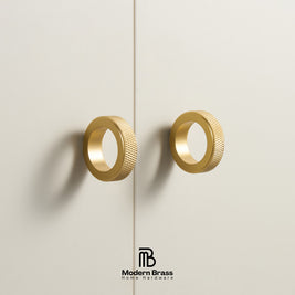 Three round cabinet knobs in black, matte brass, and brushed brass are arranged on a light beige surface against a warm beige background. The Modern Brass logo is visible at the bottom.