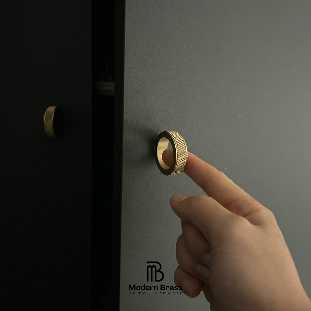 A close-up of a hand pressing a textured brass cabinet knob on a dark gray door, with a second matching brass knob visible on a black door to the left. The Modern Brass logo is printed at the bottom.