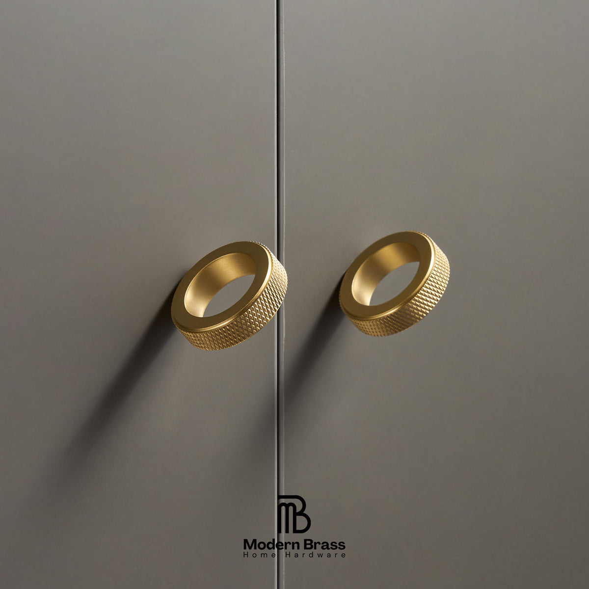 Two modern cylindrical cabinet handles in brushed gold with a knurled textured outer surface, mounted on matte dark gray doors. The Modern Brass logo is visible at the bottom.
