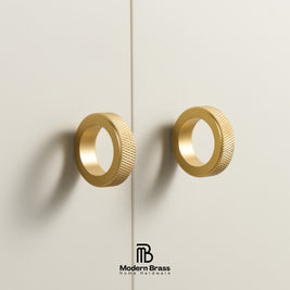 Two modern cylindrical brass cabinet pulls with a knurled textured outer edge and a polished gold interior, mounted on a light beige wall. The Modern Brass logo is visible at the bottom.