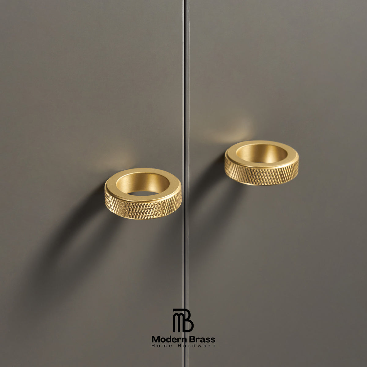 Two modern cylindrical cabinet pulls in a brushed gold finish with a knurled textured edge, mounted on a dark gray cabinet door. The Modern Brass logo is visible at the bottom.