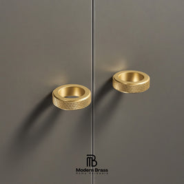 Two modern cylindrical cabinet pulls in a brushed gold finish with a knurled textured edge, mounted on a dark gray cabinet door. The Modern Brass logo is visible at the bottom.