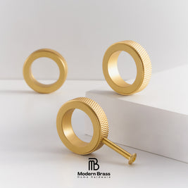 Three gold-finished brass ring hardware pieces with knurled outer edges and threaded studs, arranged on a clean white studio surface with a white pedestal. The Modern Brass Home Hardware logo appears at the bottom.
