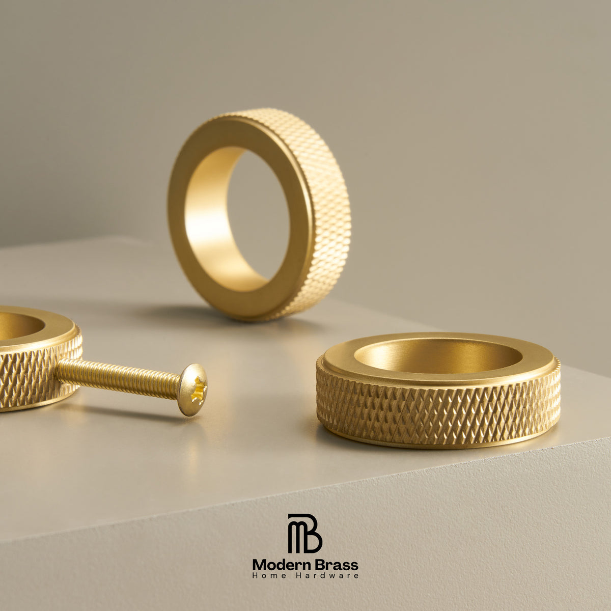 Three gold-toned textured metal ring components, including two ring bands and a threaded screw, arranged on a light gray surface with a soft neutral background. The Modern Brass Home Hardware logo appears at the bottom.