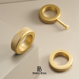 Three gold brass ring components, including a knurled band, a flat ring, and a post with a ball end, arranged on a light beige geometric background. The Modern Brass logo is visible at the bottom.