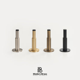 Four modern cylindrical hardware pieces in brushed silver, gold, and black finishes arranged in a row on a light gray background, with the Modern Brass Home Hardware logo below.