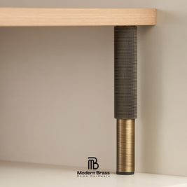 A close-up of a Modern Brass table leg featuring a textured dark gray mesh upper section and a brushed brass lower section, shown supporting a light wood shelf against a neutral beige background.
