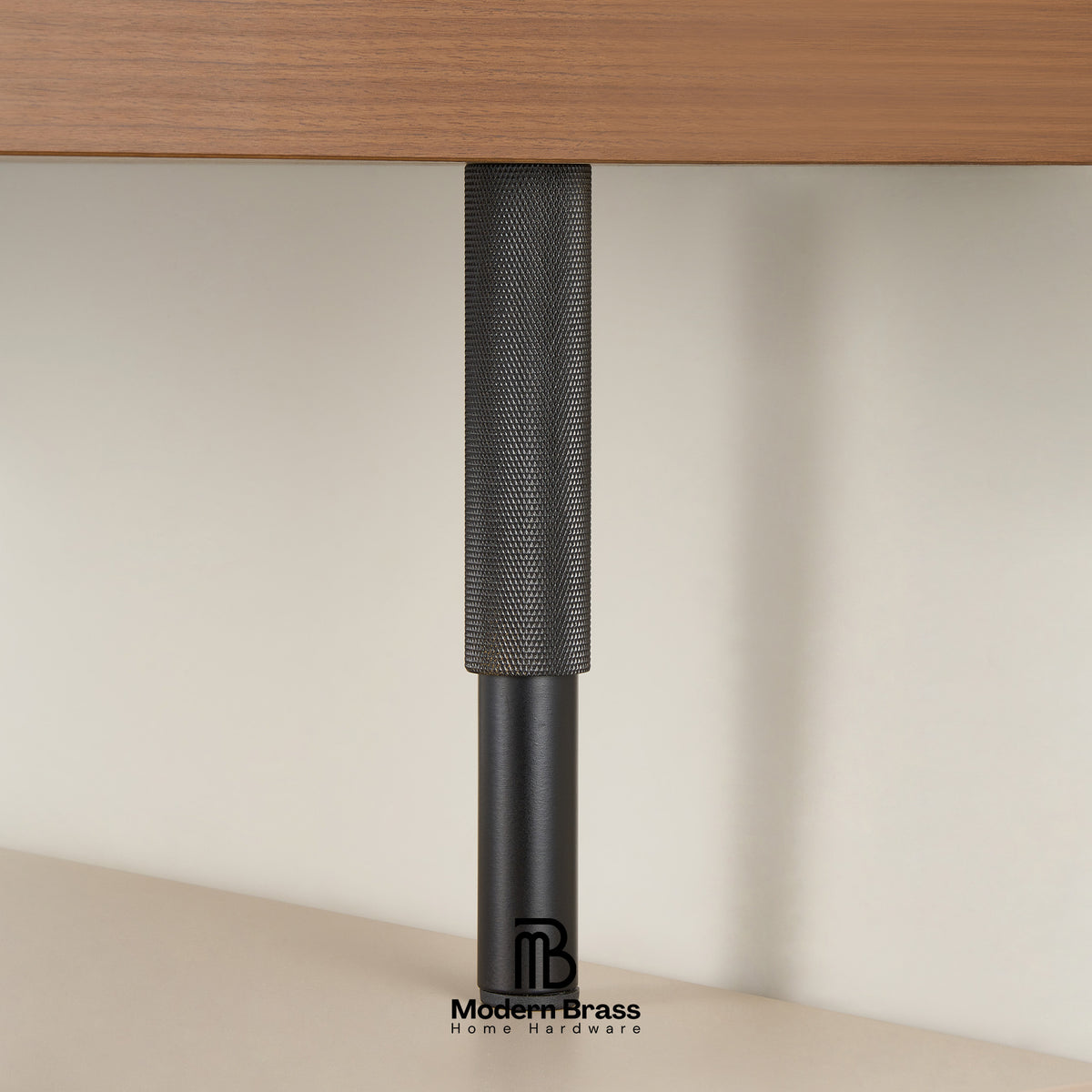 A close-up of a black metal table leg with a textured cylindrical top and a matte lower section, shown under a wooden desk surface against a light beige wall.