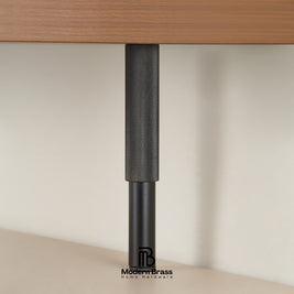 A close-up of a black metal table leg with a textured cylindrical top and a matte lower section, shown under a wooden desk surface against a light beige wall.