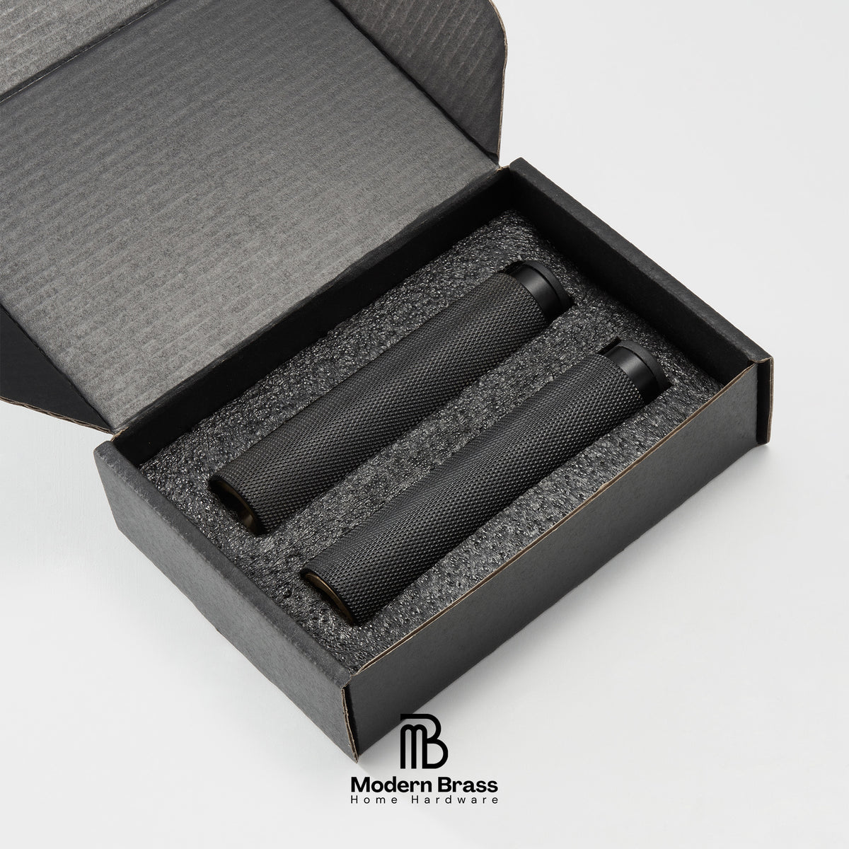Two black textured cylindrical products displayed inside an open black presentation box with foam insert, on a light gray background. The Modern Brass Home Hardware logo is visible at the bottom.