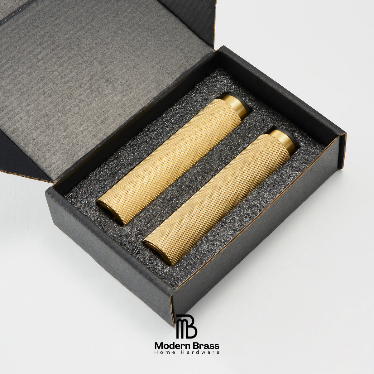 Two brass cylindrical home decor pieces displayed inside an open black presentation box with gray foam insert, on a white background. The Modern Brass logo is visible at the bottom.
