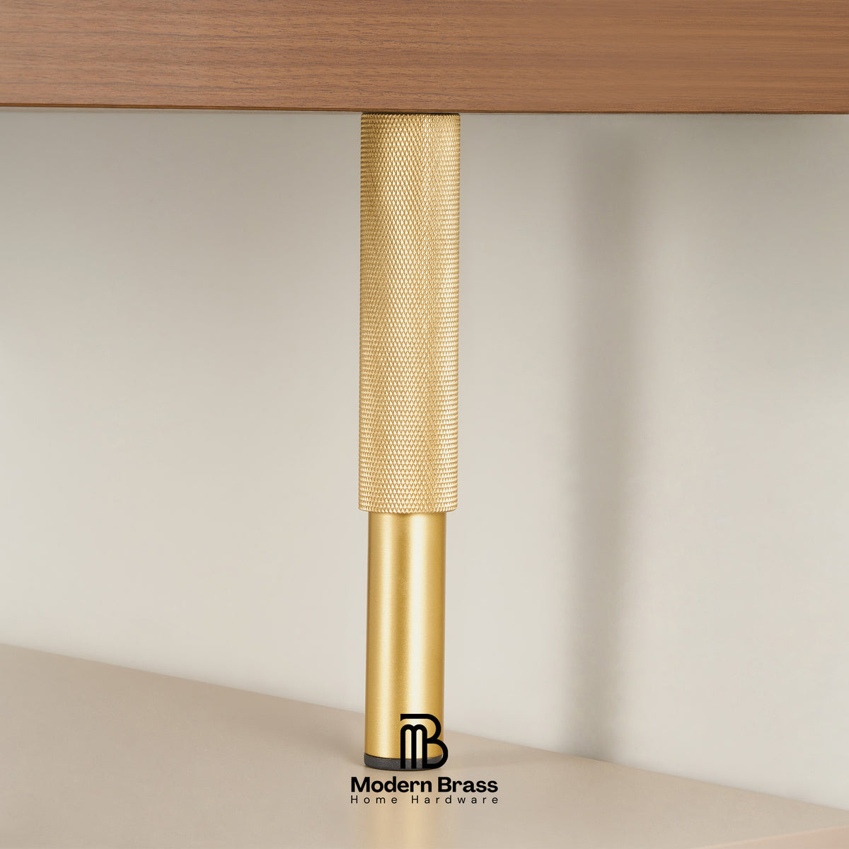 A close-up of a modern brass table leg with a textured knurled upper section and a smooth cylindrical lower section, shown supporting a wooden tabletop against a neutral beige background. The Modern Brass logo is visible at the bottom.